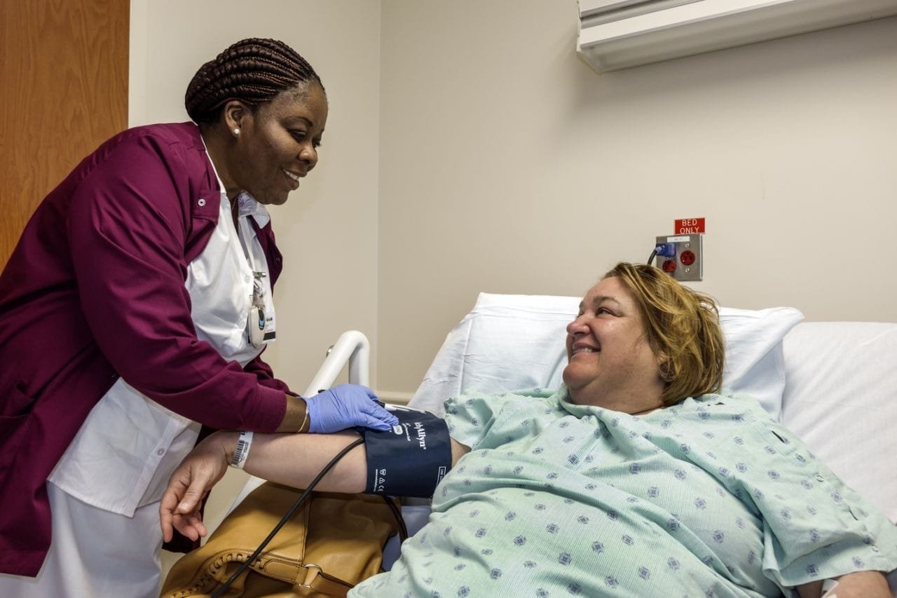 Registered nurse in burgundy scrubs and a white coat taking a patient’s blood pressure in a hospital